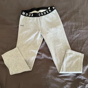 Grå leggings från Under Armour -  grå leggings från Under Armour med svart resår i midjan och vit logga. Perfekta för träning eller en avslappnad dag. De är tillverkade i ett mjukt och stretchigt material för optimal komfort.