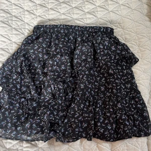 Svart blommig kjol från Stradivarius - Säljer en svart blommig kjol från Stradivarius med volanger. Kjolen har ett elastiskt midjeband och ett fint blommönster i olika färger. Perfekt för en stilfull look.