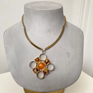 Guldhalsband med orange hänge - Unikt guldhalsband med ett stort geometriskt hänge i orange och guld. Hänget har en central rund sten omgiven av kvadratiska och cirkulära detaljer. Kedjan är i guld och har en enkel låsning.