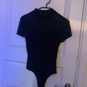 Svart bodysuit med meshdetaljer - Snygg svart bodysuit med korta ärmar och meshdetaljer upptill. Perfekt för en stilren look med en touch av elegans. Den har en hög hals och en åtsittande passform som framhäver figuren.