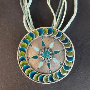 Färgglatt halsband med cirkulär hängsmycke - Unikt halsband med ett stort, cirkulärt hängsmycke i silverfärg. Smycket har ett vackert mönster med blå och gröna detaljer samt en central turkos sten. Hängsmycket är fäst vid flera tunna band i matchande färger.