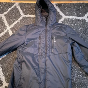 Svart jacka från Sail Racing - Snygg svart jacka från Sail Racing med huva och dragkedja. Perfekt för kyligare dagar. Jackan har flera praktiska fickor med dragkedjor och en stilren design.