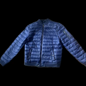 Blå dunjacka från Ralph Lauren - Säljer en snygg blå dunjacka från Ralph Lauren. Jackan har en quiltad design med dragkedja framtill och ribbade muddar. Märkets logga finns på ärmen. Perfekt för kyligare dagar!
