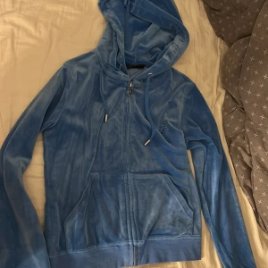 Blå hoodie från Juicy Couture - Säljer en mjuk och stilren blå hoodie från Juicy Couture. Den har en dragkedja framtill och en praktisk huva med snören. Perfekt för en avslappnad look. 💙