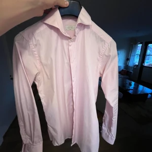 Rosa skjorta från Eton - Säljer en stilren rosa skjorta från Eton i storlek 38. Skjortan har en slim passform och är långärmad med knappar framtill. Perfekt för en elegant look. Skjortor från Eton är väldigt exklusiva pristet på just denna nu ligger på runt 1500. Denna är i bra skick inga täcken på användning.