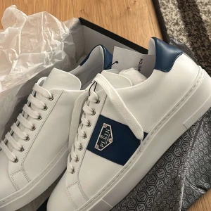 Vita sneakers med blå detaljer från Philipp Plein - Snygga vita sneakers från Philipp Plein med blå detaljer och snörning. Skorna har en rund tå och är perfekta för en stilren look. Märkesloggan syns tydligt på sidan.