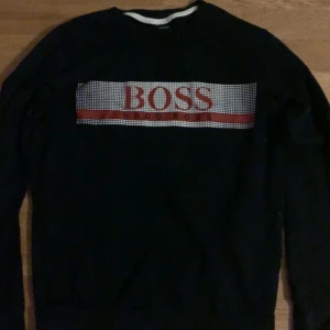 Hugo Boss tröja  - Snygg svart tröja från Hugo Boss med ett rött och vitt mönster över bröstet där märkesnamnet står. Perfekt för en stilren look.