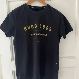 Svart t-shirt från Hugo Boss - Snygg svart t-shirt från Hugo Boss med guldtext på framsidan. Perfekt för en stilren och avslappnad look. T-shirten har en normal passform och är tillverkad i ett mjukt material.
