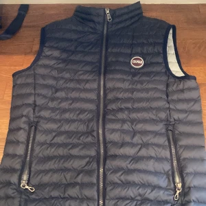 Blå dunväst från Colmar - Säljer en snygg blå dunväst från Colmar med dragkedja och två praktiska fickor framtill. Västen har en quiltad design och en hög krage för extra värme. Perfekt för kyliga dagar!
