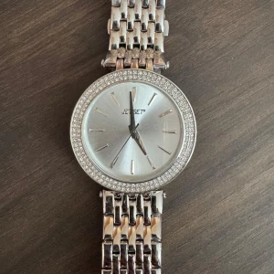 Silverfärgad klocka från Jet Set - Snygg silverfärgad klocka från Jet Set med glittrande stenar runt urtavlan. Klockan har en stilren design med metallarmband och en tydlig urtavla. Perfekt för att lägga till lite bling till din outfit.