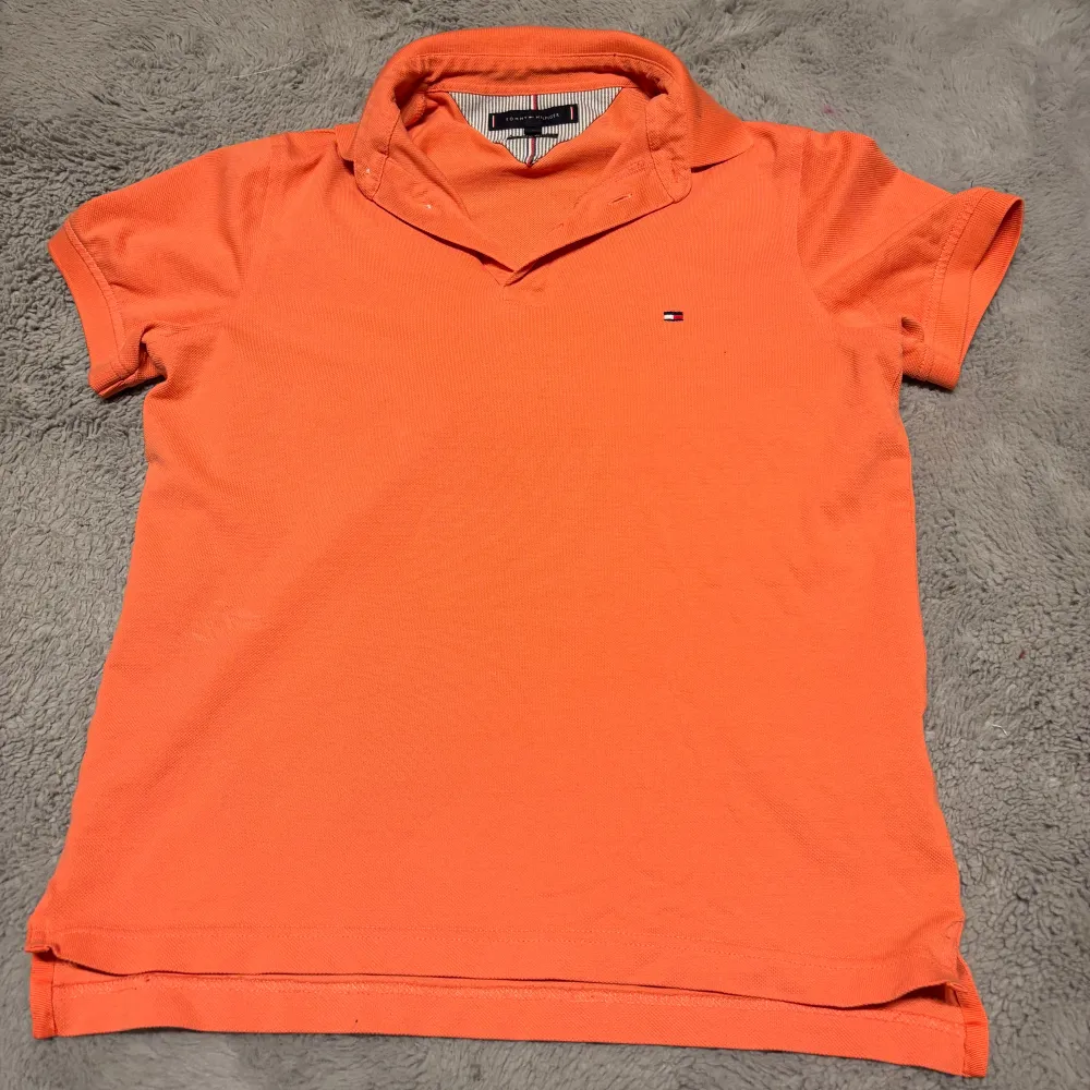 Snygg orange pikétröja från Tommy Hilfiger med klassisk krage och korta ärmar. Tröjan har en liten broderad logotyp på bröstet och är perfekt för en avslappnad stil.. T-paidat.