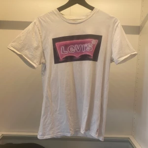Vit t-shirt från Levi's - Säljer en vit t-shirt från Levi's med ett coolt tryck i rosa och svart på bröstet. T-shirten är kortärmad och tillverkad i bomull.