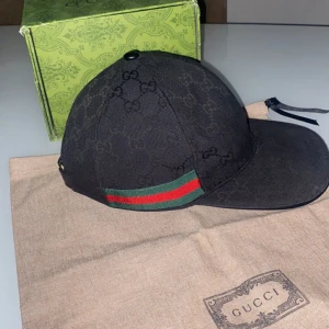 Svart keps från Gucci - Snygg svart keps från Gucci med det ikoniska GG-mönstret. Kepsen har en justerbar rem baktill och en grön och röd rand på sidan. Perfekt för att ge din outfit en lyxig touch.