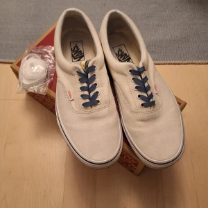 Vita Vans med blå snörning - Säljer ett par klassiska Vans Era i vit färg med blå snörning. Skorna har en unik design på sidan och kommer med extra vita skosnören. Perfekta för en stilren look. (Du kan dra white vans skämtet) använda 3ggr ish