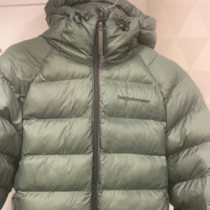 Grön dunjacka från Peak Performance - Säljer en grön dunjacka från Peak Performance med huva och dragkedja framtill. Jackan är perfekt för kyliga dagar och har en stilren design med märkets logga på bröstet. 