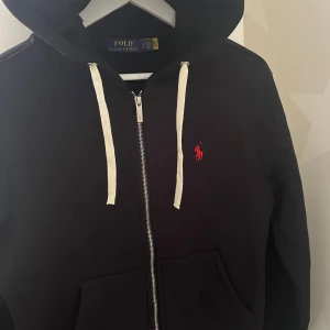 Svart hoodie från Ralph Lauren - Säljer en stilren svart hoodie från Ralph Lauren med dragkedja och vit snörning i huvan. Den har en röd logotyp på bröstet och är perfekt för en avslappnad stil. Passar bra till både jeans och joggers.
