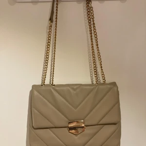 Beige quiltad handväska med kedja - Snygg beige handväska med quiltat mönster och guldfärgad kedja. Väskan har en elegant låsdetalj framtill och är perfekt för att ge en stilren touch till din outfit.