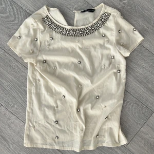 Vit blus med pärldetaljer från Dorothy Perkins - Säljer en elegant vit blus från Dorothy Perkins med korta ärmar och vackra pärldetaljer runt halsringningen. Blusen har även små pärlor utspridda över hela tyget för en extra touch av glamour. Perfekt för speciella tillfällen!