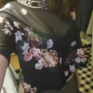 Svart blommig t-shirt från SikSilk - Säljer en svart t-shirt från SikSilk med vackert blommönster i rosa och vita nyanser. T-shirten har korta ärmar och en rund halsringning med märkets namn tryckt runtom. Perfekt för en stilren och trendig look.