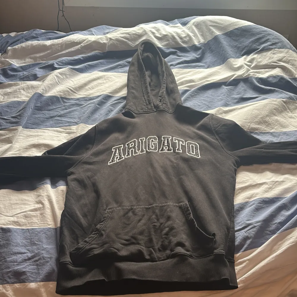 Säljer en svart hoodie från Axel Arigato med texten 'Arigato' i vitt på framsidan. Den har en klassisk känguruficka och en bekväm huva. Perfekt för en avslappnad stil. Det är några små hål framme på tröjan . Neuletakit & Villapaidat.