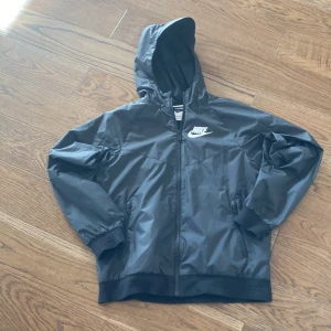 Svart vindjacka från Nike - Säljer en stilren svart vindjacka från Nike med huva och dragkedja. Jackan har långa ärmar och ribbade muddar vid ärmslut och nederkant. Perfekt för blåsiga dagar!