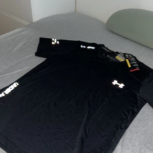 Reflective Under Armour - Snygg svart t-shirt från Under Armour med logga på bröstet och textdetaljer på ärmarna. Perfekt för träning eller en avslappnad dag. Tillverkad i ett lätt och andningsbart material.