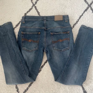 Snygga blåa Nudie Jeans - Snygga blå jeans från Nudie Jeans med klassisk femficksdesign och orange sömmar. Jeansen har en normal passform och är perfekta för en avslappnad stil. De har en knappgylf och är tillverkade i slitstarkt denim.