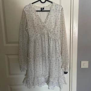 Vit blommig klänning med volangdetaljer - Charmig vit klänning med små blå blommor och volangdetaljer. Klänningen har en v-ringad design och långa ärmar, perfekt för vår och sommar. Den har en omlott-look och är gjord i ett lätt och luftigt material.