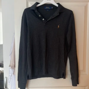Grå pikétröja från Ralph Lauren - Säljer en stilren grå pikétröja från Ralph Lauren med broderad logga på bröstet. Tröjan har långa ärmar och en klassisk krage med knappar. Perfekt för en avslappnad men ändå elegant look.