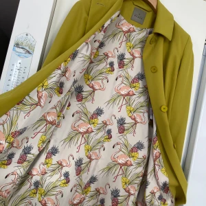 Olivgrön kappa från Fuchs Schmitt.size 38.ny - Elegant olivgrön kappa från Fuchs Schmitt med ett färgglatt foder prytt av flamingos och ananas. Kappan har en klassisk krage och knäpps med knappar framtill. Den har ett bälte i midjan för en smickrande passform.