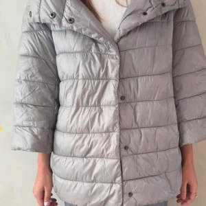 Grå pufferjacka med trekvartsärm - Snygg och stilren grå pufferjacka med trekvartsärm. Jackan har en quiltad design och knappar framtill. 