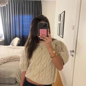 Beige stickad tröja från Gina Tricot - Säljer en snygg beige stickad tröja från Gina Tricot med puffiga korta ärmar och ett flätat mönster. Perfekt för en stilren look. Tröjan har en rund halsringning och ribbade kanter.