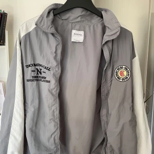 Grå och vit vindjacka från Bershka - Snygg grå och vit vindjacka från Bershka med dragkedja framtill. Jackan har tryck på bröstet och en broderad patch på sidan. Perfekt för en sportig look. utan defekter. Vill du ha fler bilder så skriv.