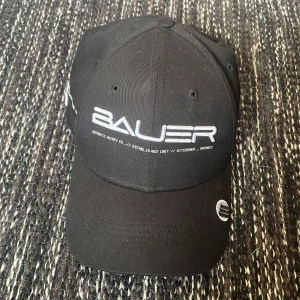 Svart keps från Bauer - Snygg svart keps från Bauer med broderad logga framtill och textdetaljer på sidorna och baksidan. Justerbar passform med spänne bak. Perfekt för hockeyfans! Oanvänd men namnmärkt inuti kepsen. 