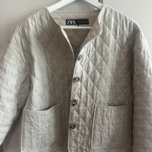 Beige quiltad jacka från Zara - Säljer en beige quiltad jacka från Zara med knappar framtill. Jackan har en rund halsringning och stora fickor på framsidan. Perfekt för en stilren look.