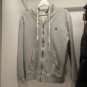 Grå hoodie från Polo Ralph Lauren - Säljer en stilren grå hoodie från Polo Ralph Lauren med dragkedja och justerbar huva. Tröjan har långa ärmar och en broderad logotyp i blått på bröstet. Perfekt för en avslappnad look.