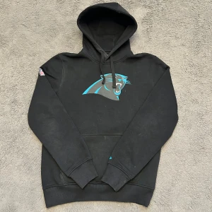 New era svart hoodie  - Svart hoodie med Carolina Panthers logga tryckt på framsidan. Hoodien har en ficka på magen. NFL loggan finns tryckt på ena ärmen och New Era loggan på den andra. Storlek M