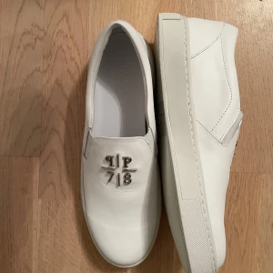 Vita loafers från Philipp Plein - Snygga vita loafers från Philipp Plein med en stilren design och metallisk logga på ovansidan. Skorna har en rund tå och är tillverkade i skinn, vilket ger en lyxig känsla. Perfekta för en stilren look.