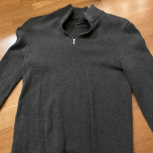 Mörkgrå halfzip - Snygg mörkgrå tröja från Ados Design med dragkedja vid halsen. Tröjan har en ribbad krage och är perfekt för våren! Den är långärmad och har en stilren design som passar till många tillfällen. Passar också xxs