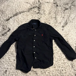 Svart skjorta från Ralph Lauren - Säljer en stilren svart skjorta från Ralph Lauren med klassisk krage och knappar framtill. Skjortan har en broderad logotyp i rött på bröstet och långa ärmar. Självklart äkta. Storlek S, passar folk rund 170-180. Skriv vid funderingar!