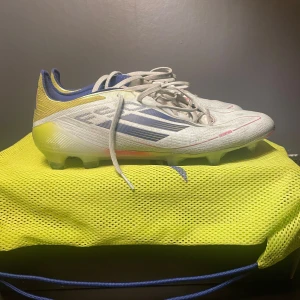  fotbollsskor från Adidas - Snygga vita fotbollsskor från Adidas med blå och gula detaljer. Skorna har snörning och en rund tå, perfekt för fotbollsplanen. De kommer med en praktisk gul förvaringspåse.