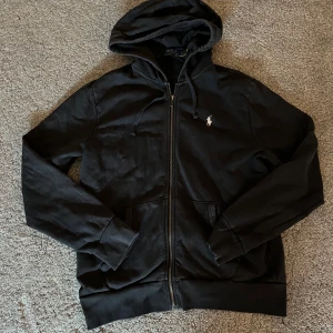 Svart hoodie från Polo Ralph Lauren - Säljer en stilren mörkblå hoodie från Polo Ralph Lauren med dragkedja och en liten broderad logga på bröstet. Perfekt för en avslappnad look. Den har långa ärmar och en praktisk huva med dragsko.