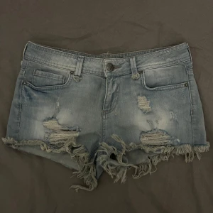 Lågmidjade jeansshorts  - Snygga blå lågmidjade jeansshorts med slitna detaljer och fransar vid benen. Perfekta för en avslappnad sommarlook. De har en klassisk femficksdesign och knappgylf.