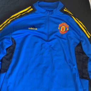 Blå träningsjacka från Adidas - Säljer en blå träningsjacka från Adidas med Manchester United-logga på bröstet. Jackan har gula ränder längs ärmarna och en dragkedja vid halsen. Perfekt för fotbollsträning eller som en sportig vardagslook.