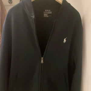 Mörkblå hoodie från Polo Ralph Lauren - Snygg svart hoodie från Polo Ralph Lauren med dragkedja och det klassiska logotypbroderiet på bröstet. Perfekt för en stilren och avslappnad look.