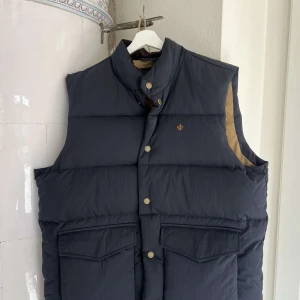 Mörkblå dunväst från Morris - Säljer en snygg mörkblå dunväst från Morris med stilrena knappar och en liten broderad logga på bröstet. Västen har en hög krage och praktiska fickor framtill. Perfekt för kyliga dagar!