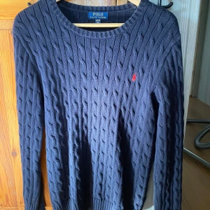 Mörkblå stickad tröja från Polo Ralph Lauren - Säljer en stilren mörkblå stickad tröja från Polo Ralph Lauren med kabelstickat mönster. Tröjan har långa ärmar och en liten röd logotyp på bröstet. Perfekt för kyligare dagar.