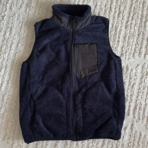 Mörkblå fleeceväst - Snygg mörkblå fleeceväst med dragkedja och hög krage. Västen har en praktisk bröstficka med dragkedja och är perfekt för kyliga dagar. Den är både stilren och funktionell.