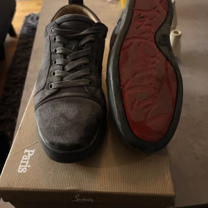 Grå sneakers från Christian Louboutin - Snygga grå sneakers från Christian Louboutin med den ikoniska röda sulan. Skorna har snörning och en rund tå, vilket ger en klassisk och stilren look. Perfekta för den som vill ha en touch av lyx i vardagen. På bild 5 ser ni att två knappar saknas därav priset🤗 (pris kan diskuteras)