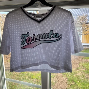 Vit croppad mesh-topp från FB Sister - Säljer en vit croppad mesh-topp från FB Sister med texten 'Toronto' i färgglada bokstäver. Toppen har en v-ringad hals och korta ärmar, perfekt för en sportig look. 🏀